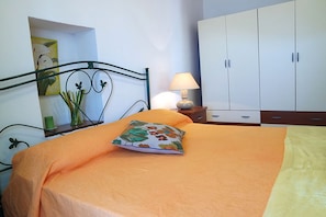 A neatly made bed with an orange bedspread and a floral pillow, a bedside lamp, a green vase with plants, and a white wardrobe. - Bella Mbriana Apartments (Lustra)