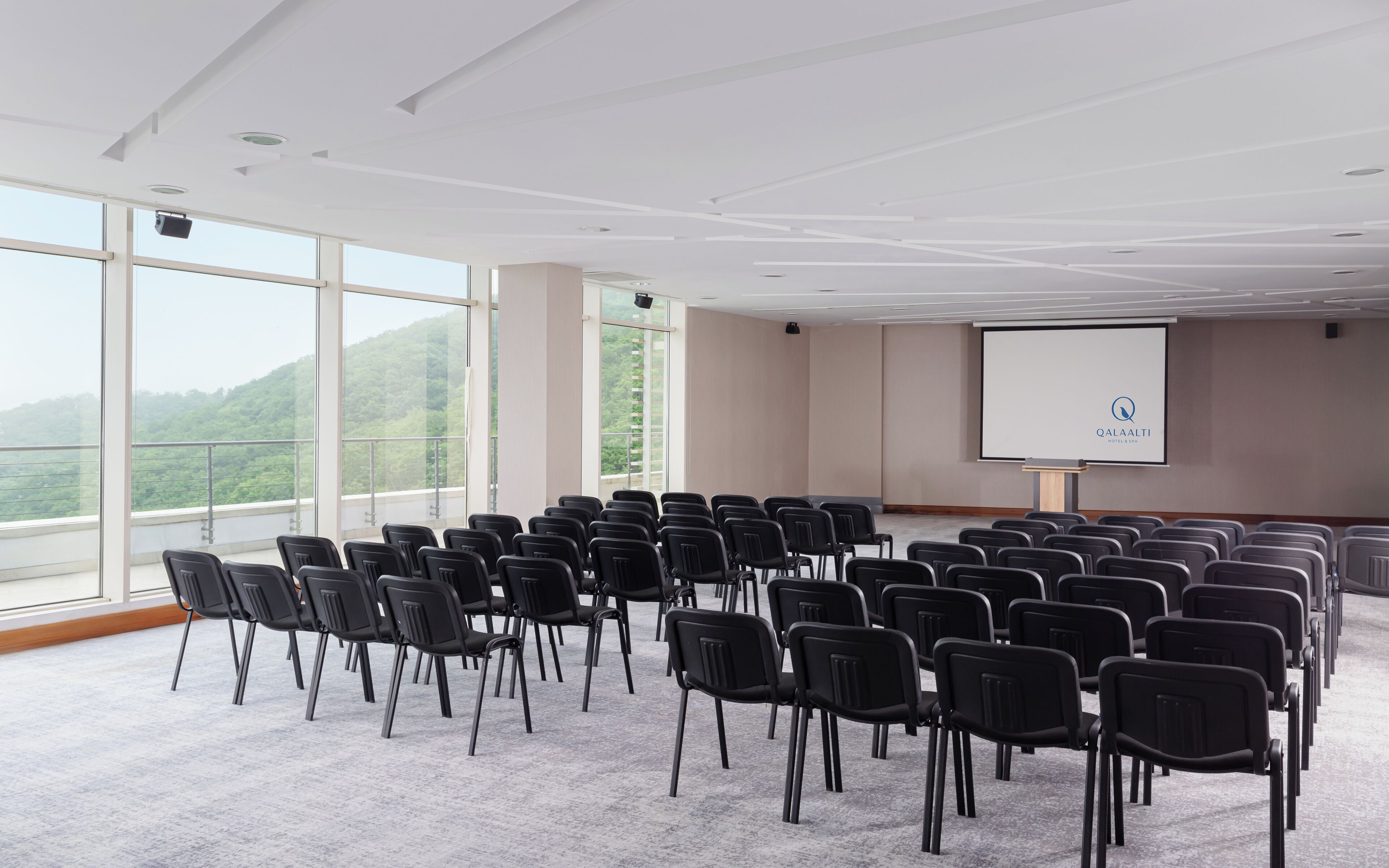 A conference room with rows of black chairs, a projector screen, and large windows offering a view of green hills.