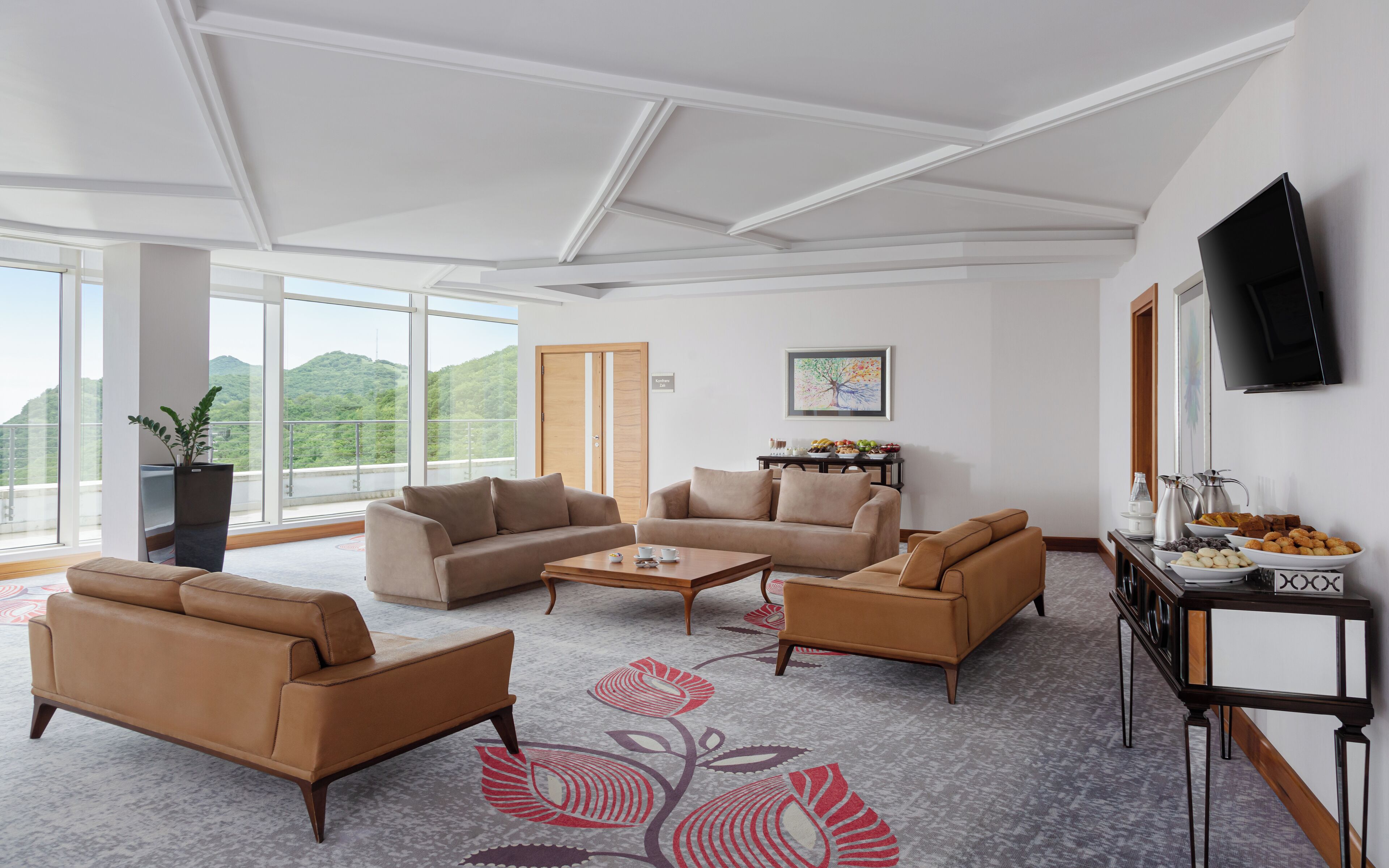 A spacious living room with a large sectional sofa, a coffee table, and a TV mounted on the wall. There is a balcony with a view of green hills.