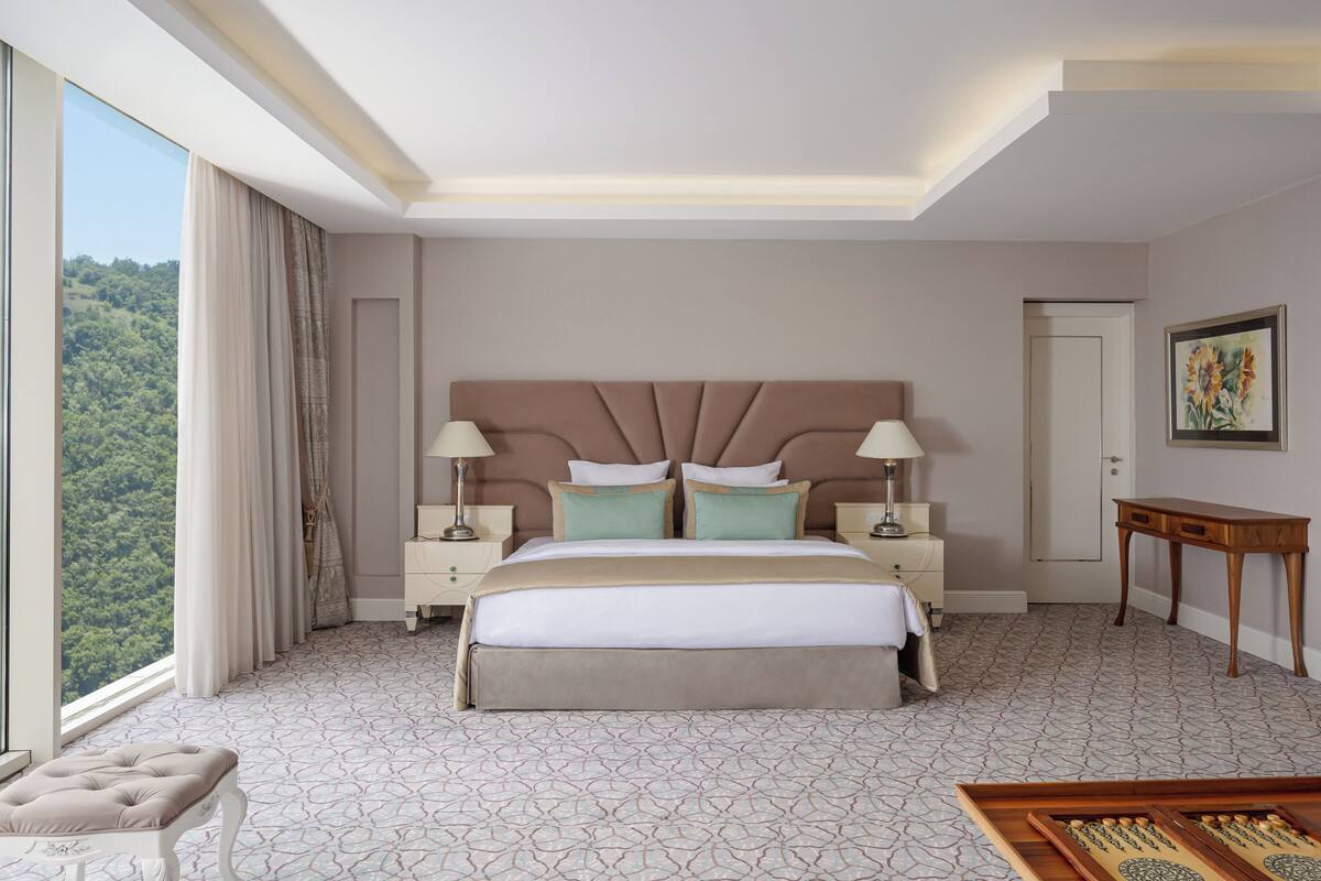 A hotel room with a large bed, two bedside tables, a nightstand, a wooden side table, a view of greenery through the window, and a painting on the wall.