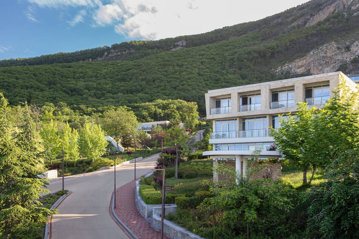A modern building with a stone facade and multiple balconies, surrounded by lush greenery and a winding road.