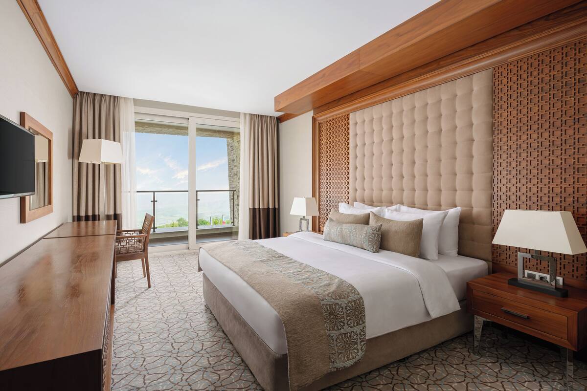 A hotel room with a large bed, a desk, a TV, and a balcony with a view.