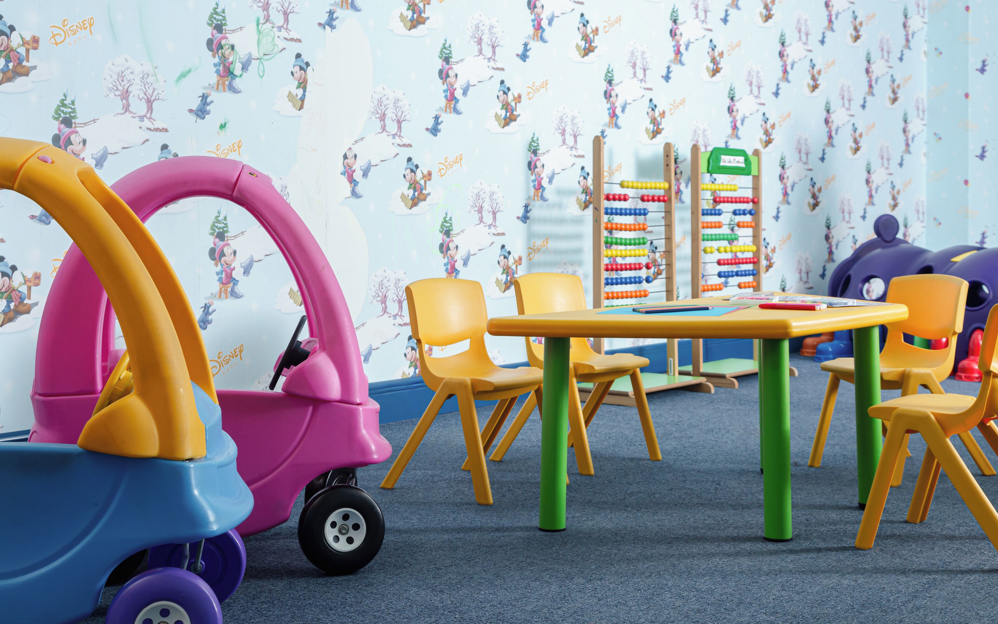 A children's play area with colorful toys, a table, chairs, and a wall with Disney-themed wallpaper.