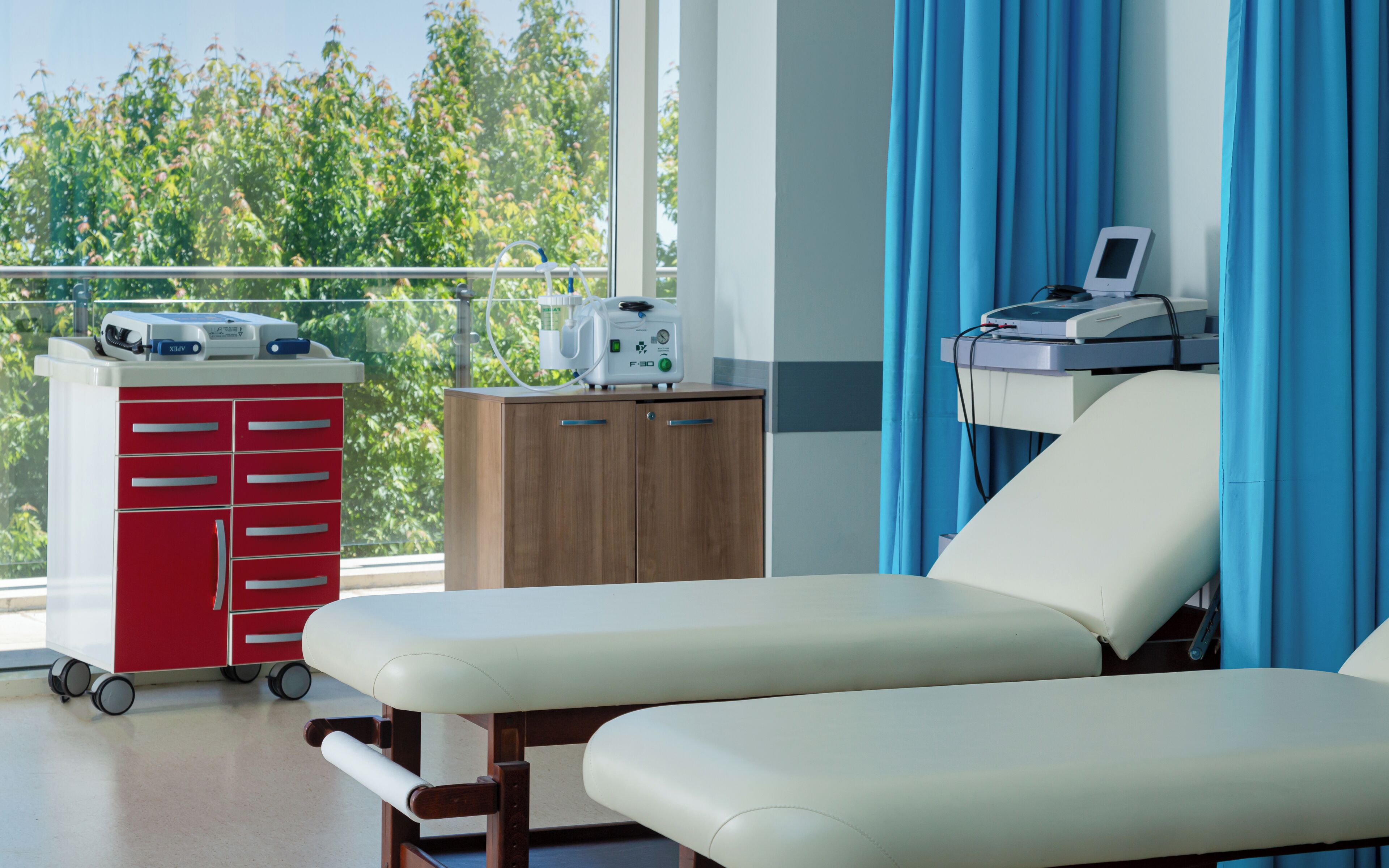 A medical examination room with two examination beds, a red medical cabinet, and a window with a view of trees.