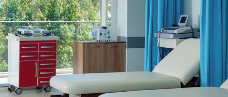 A medical examination room with two examination beds, a red medical cabinet, and a window with a view of trees.