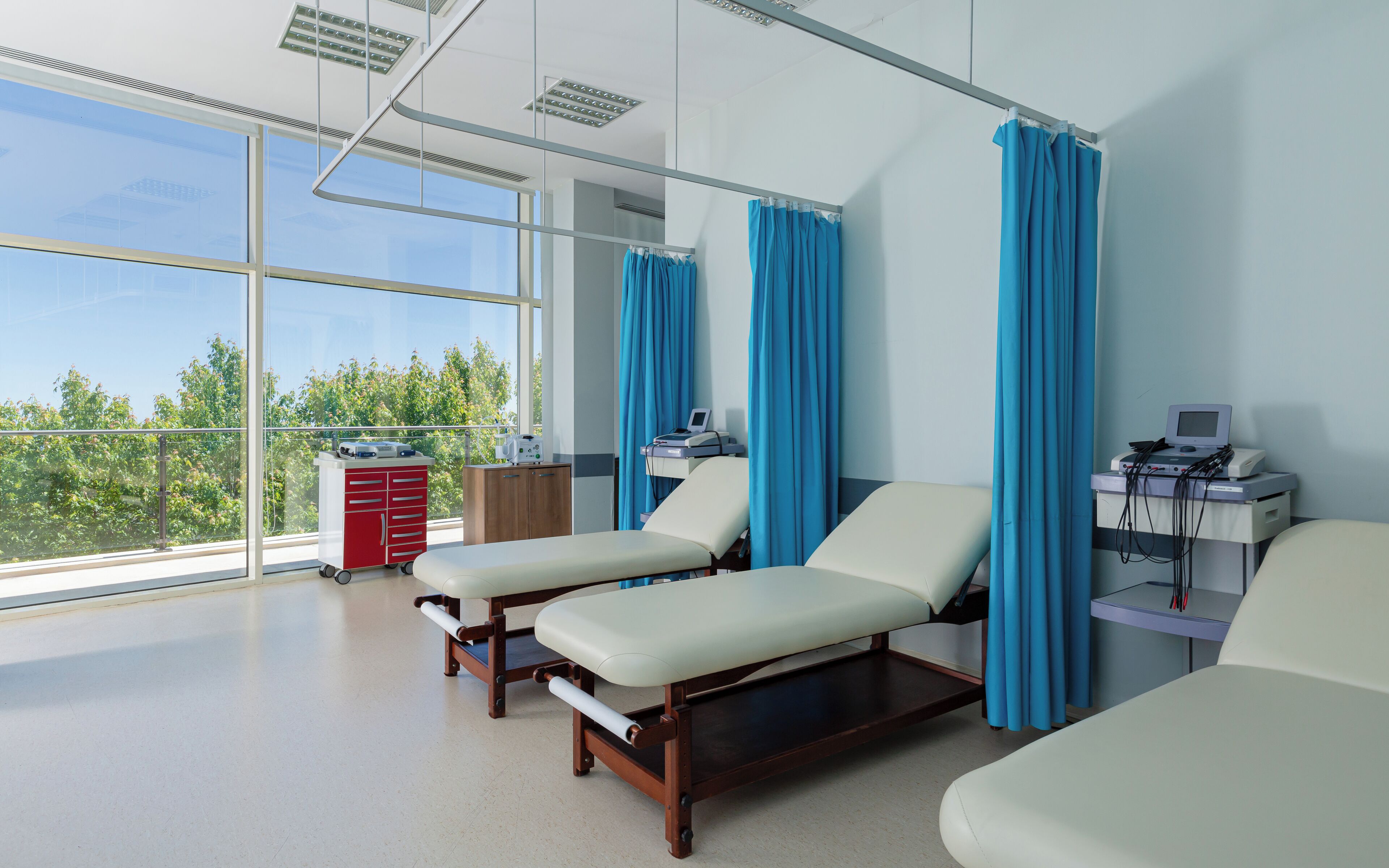A medical examination room with two examination beds, a medical cart, and a window with a view of trees.
