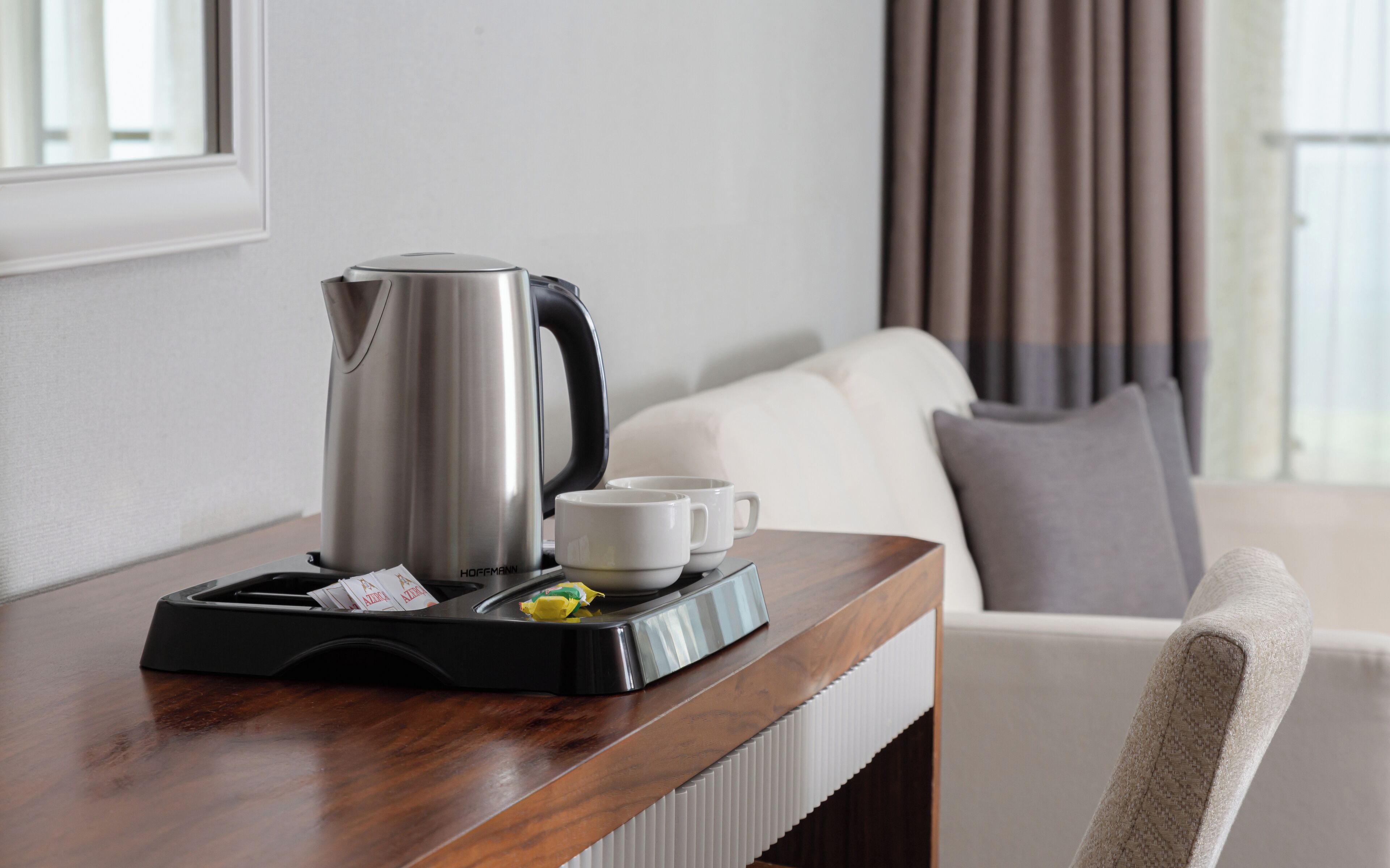 A hotel room with a wooden side table holding a silver electric kettle, a white cup, and a small yellow item.