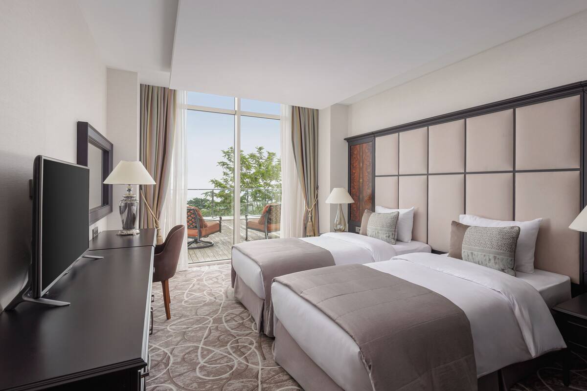 A hotel room with two beds, a desk with a TV, and a view of the outdoors.