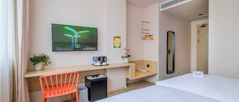 A hotel room with a bed, a desk with a chair, a TV, a small trash bin, a potted plant, and a mirror.