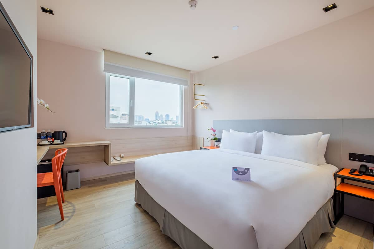 Une chambre d’hôtel avec un grand lit, un bureau avec une chaise, une petite table avec un téléphone et une fenêtre donnant sur la ville.