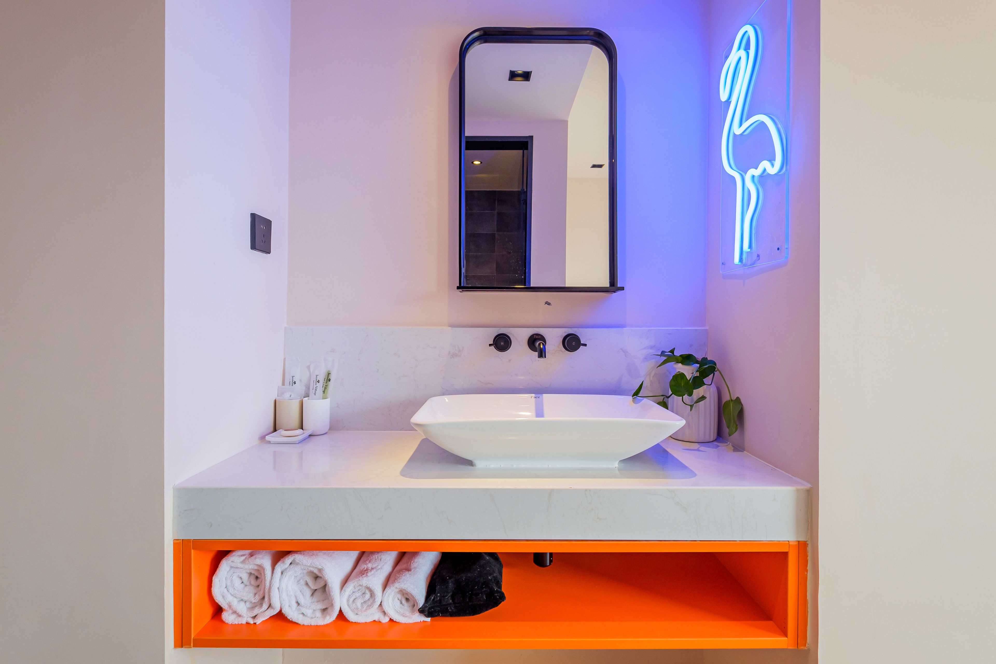 Une salle de bain avec un lavabo blanc, un miroir et un néon en forme de flamant rose.