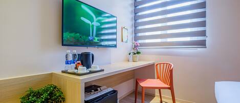 A modern hotel room with a flat-screen TV mounted on the wall, a small desk with a coffee maker, a refrigerator, a potted plant, and a window with blinds.