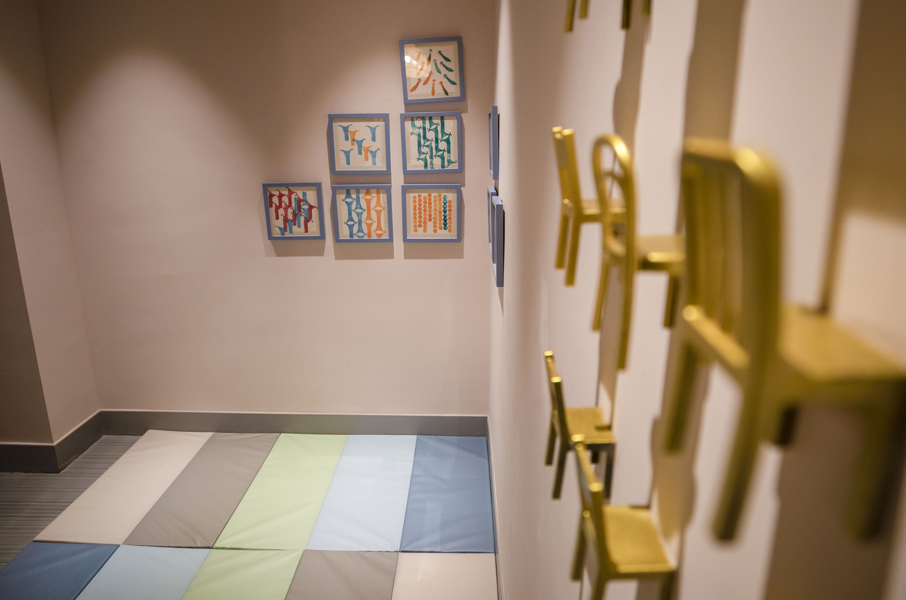 A hallway with a patterned floor, a wall with framed artwork, and a row of golden chairs.