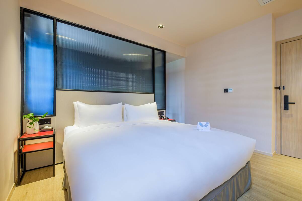 Une chambre d’hôtel moderne avec un grand lit, une table de chevet agrémentée d’une plante et une grande fenêtre avec des stores.