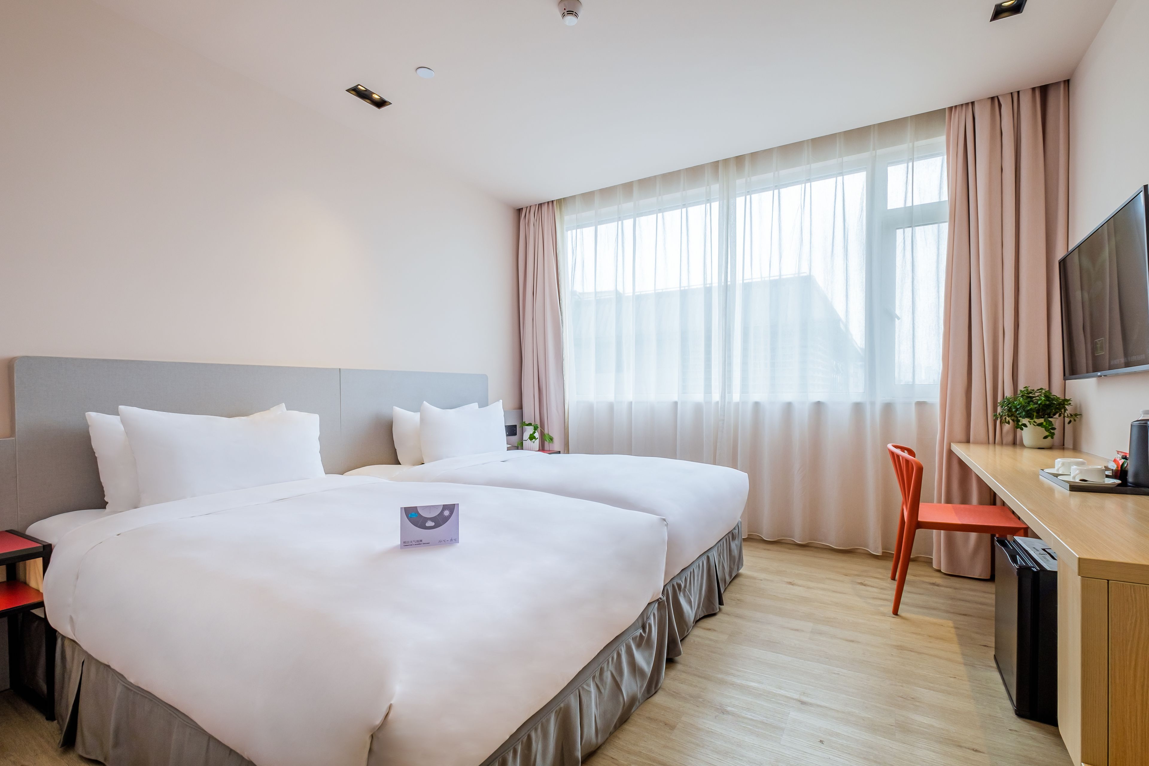 Habitación de hotel con dos camas, un escritorio, un televisor y una ventana con cortinas translúcidas.