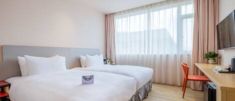 A hotel room with two beds, a desk, a TV, and a window with sheer curtains.