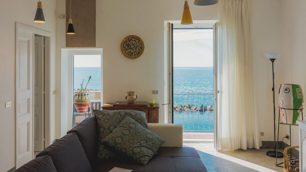 A living room with a sofa, a view of the sea through a sliding door, and pendant lights. - La Concha Suites (Pollica)
