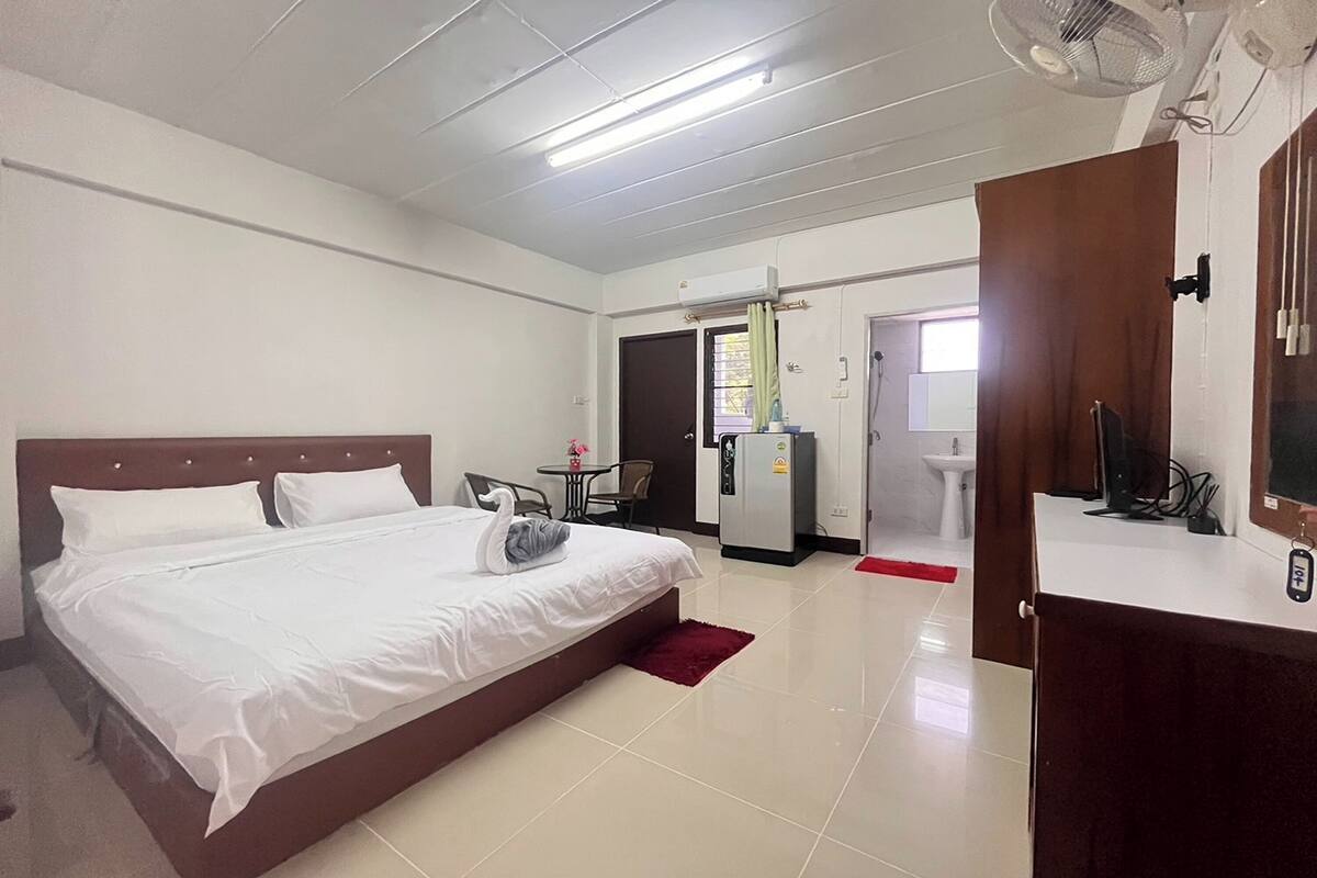Une chambre d’hôtel avec un grand lit, un bureau avec un ordinateur, un appareil de climatisation et une salle de bain visible à travers une porte ouverte.