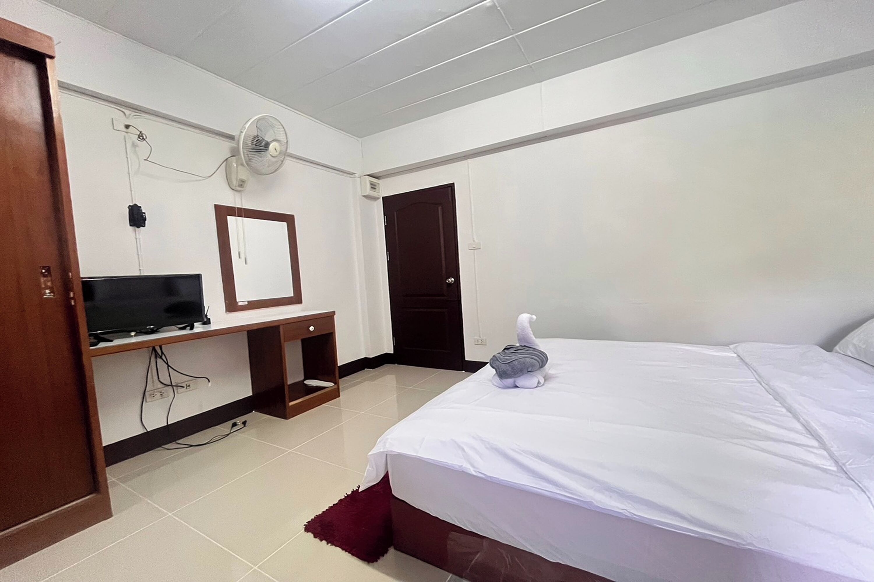 Ein Hotelzimmer mit einem Bett, einem Schreibtisch mit Fernseher, einem Wandventilator und einer Tür.