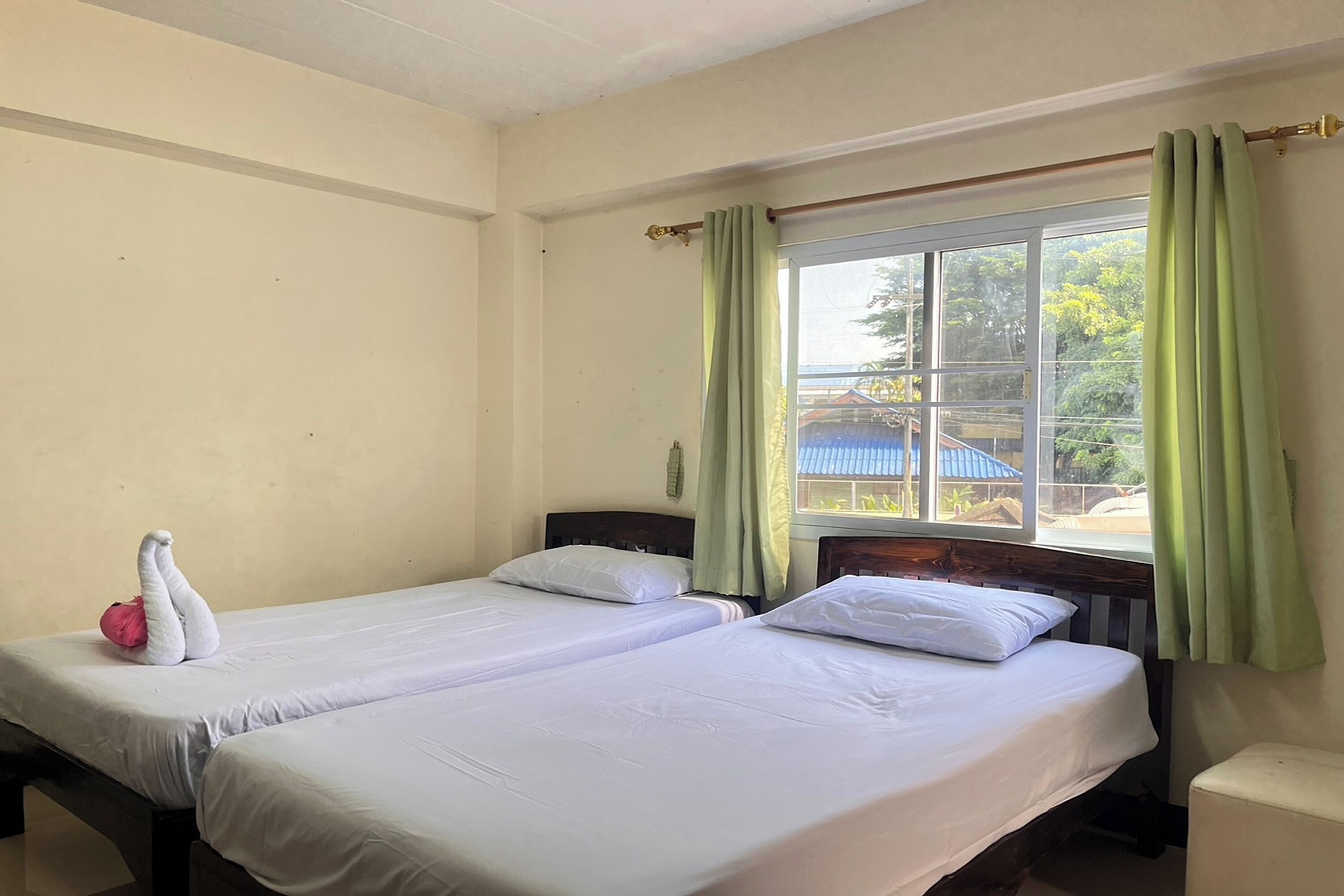 A room with two single beds, a window with green curtains, and a view of an outdoor area with a blue building and trees.