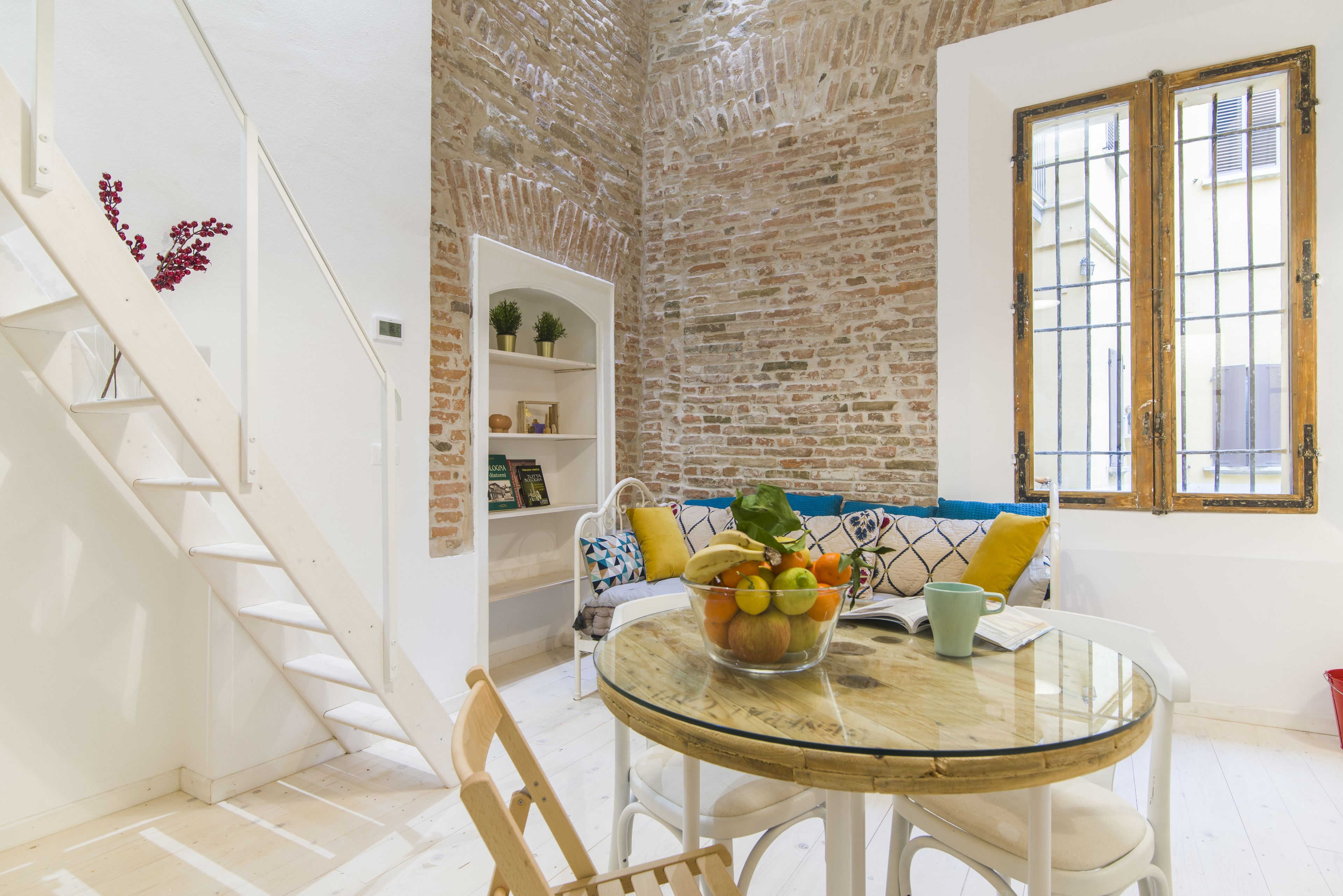 Un área de comedor con una mesa de vidrio, sillas blancas, una pared de ladrillo, una escalera blanca y una estantería con objetos decorativos.