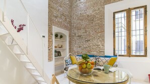 A dining area with a glass table, white chairs, a brick wall, a white staircase, and a bookshelf with decorative items. - University Loft Apartment (Bologna)
