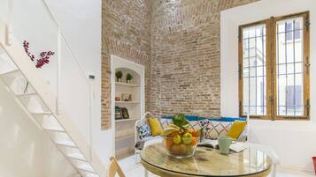 Un área de comedor con una mesa de vidrio, sillas blancas, una pared de ladrillo, una escalera blanca y una estantería con objetos decorativos.