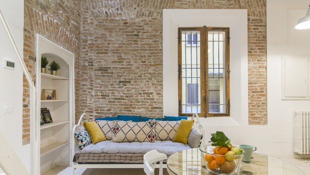 A cozy living room with a brick wall, a sofa with patterned cushions, a glass table with fruit, and a window with shutters. - University Loft Apartment (Bologna)