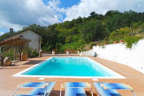 Una piscina rettangolare con acqua cristallina, circondata da un pontile in legno. Sono disposte poltrone prendisole con cuscini blu di fronte alla piscina.