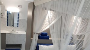 A bedroom with a bed, a bedside table, a mirror, and a bathroom with a sink and mirror. - Serenity Nhabanga (Praia do Bilene)