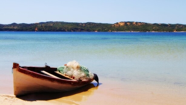 A wooden boat on a sandy beach with clear water and a hilly landscape in the background. - Serenity Nhabanga (Praia do Bilene)