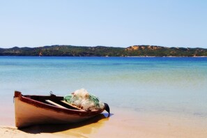 A wooden boat on a sandy beach with clear water and a hilly landscape in the background. - Serenity Nhabanga (Praia do Bilene)