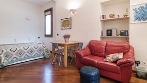 Una habitación con una cama, un sofá de cuero rojo, una mesa de comedor de madera con sillas, una mesita con una cesta y un estante de pared con adornos.