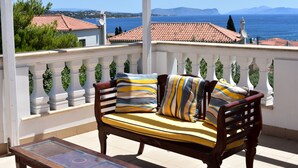 Un balcon doté d’un banc en bois et de coussins, offrant une vue sur la côte, avec des bâtiments et des montagnes en toile de fond.
