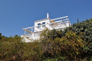 A white building with a balcony and a tower, surrounded by dense foliage.