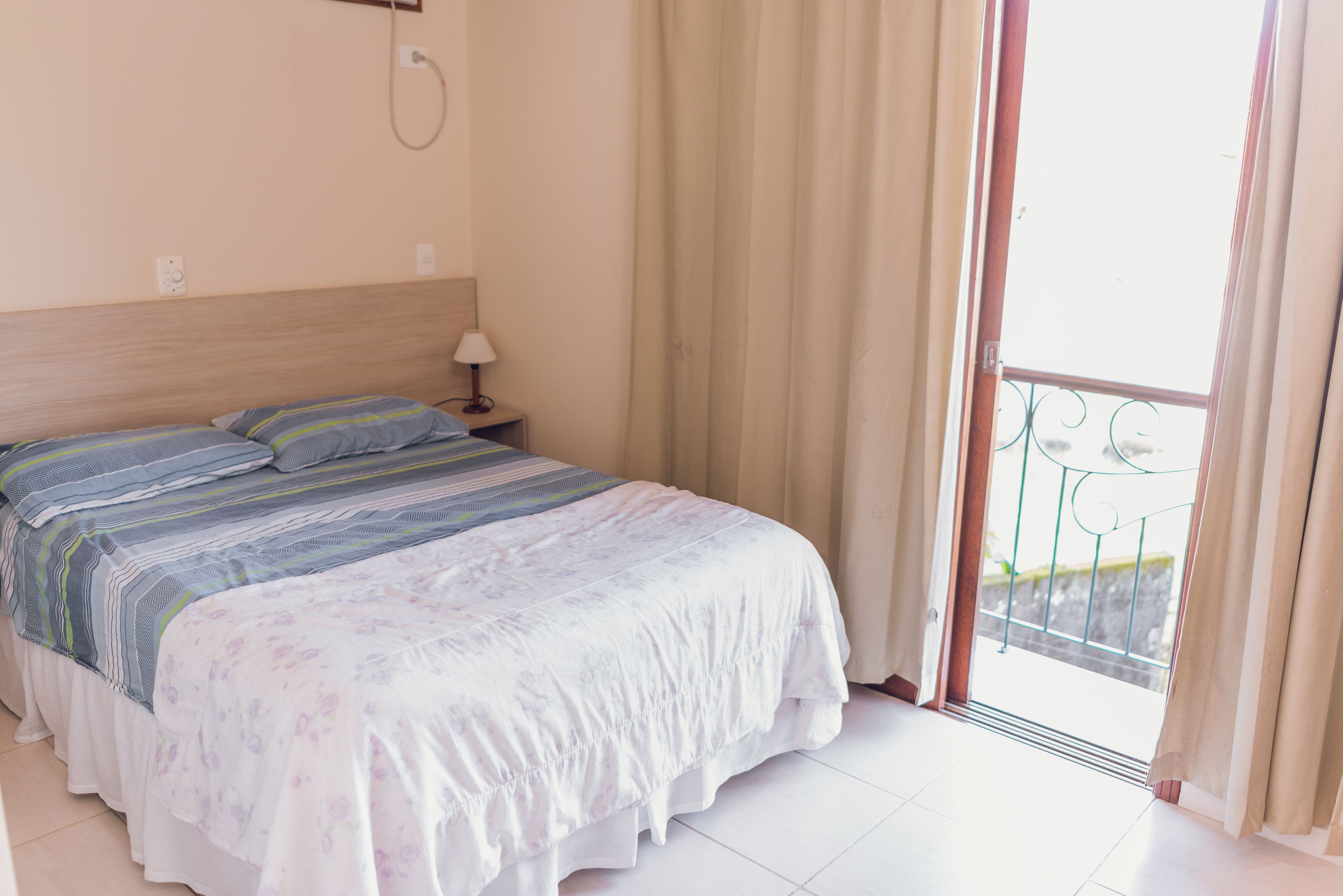 Habitación de hotel con cama, mesita de noche, lámpara y vistas al balcón exterior.