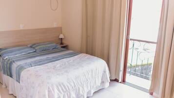 Habitación de hotel con cama, mesita de noche, lámpara y vistas al balcón exterior.