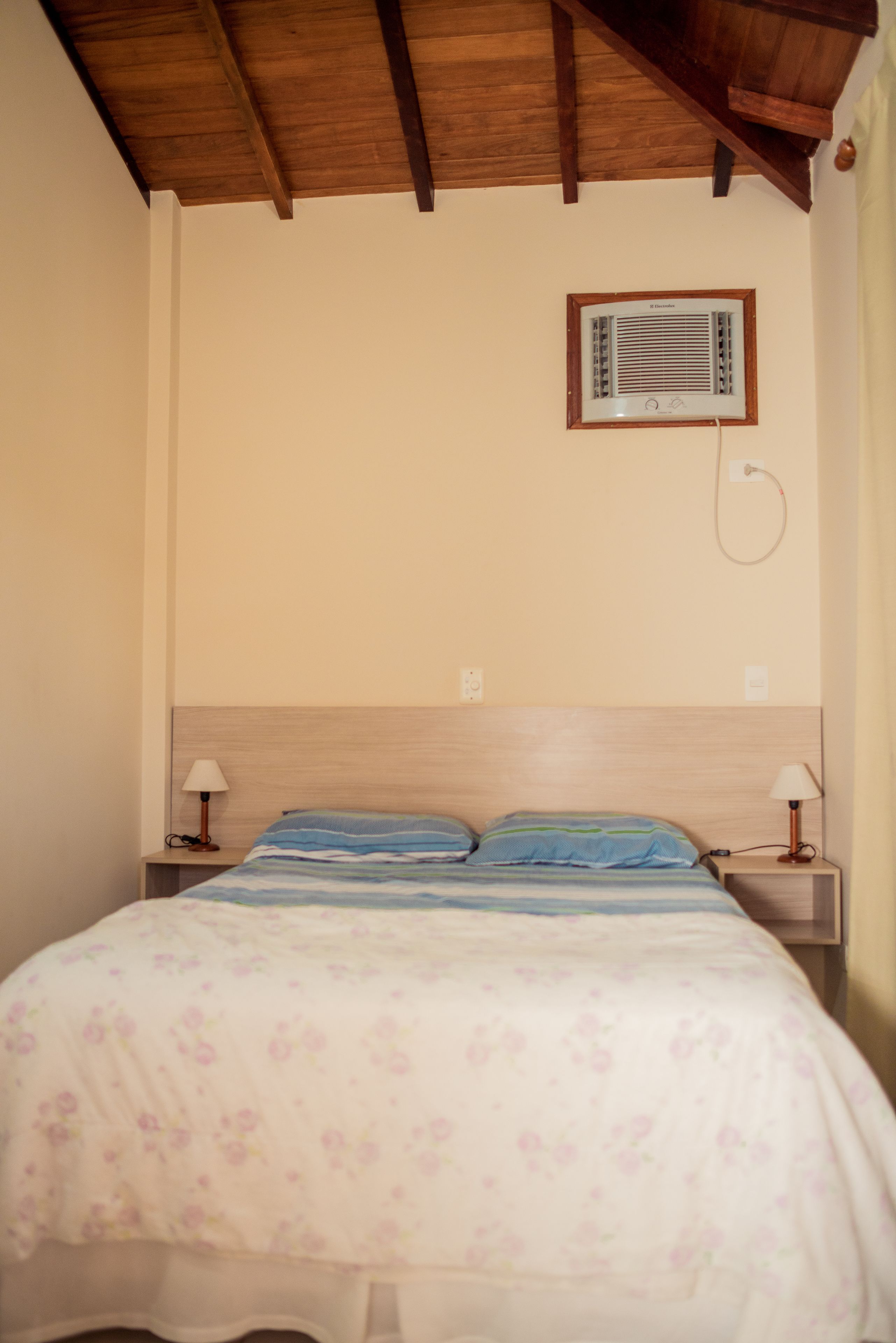 Quarto com teto de madeira, cama com roupa de cama floral, duas luminárias de mesa e aquecedor fixado na parede.