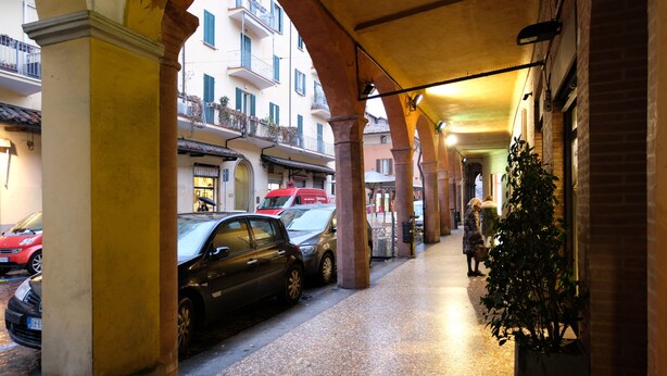 A covered pedestrian walkway with a row of parked cars and a building with balconies. - Castiglione Lovely Mansarda (Bologna)