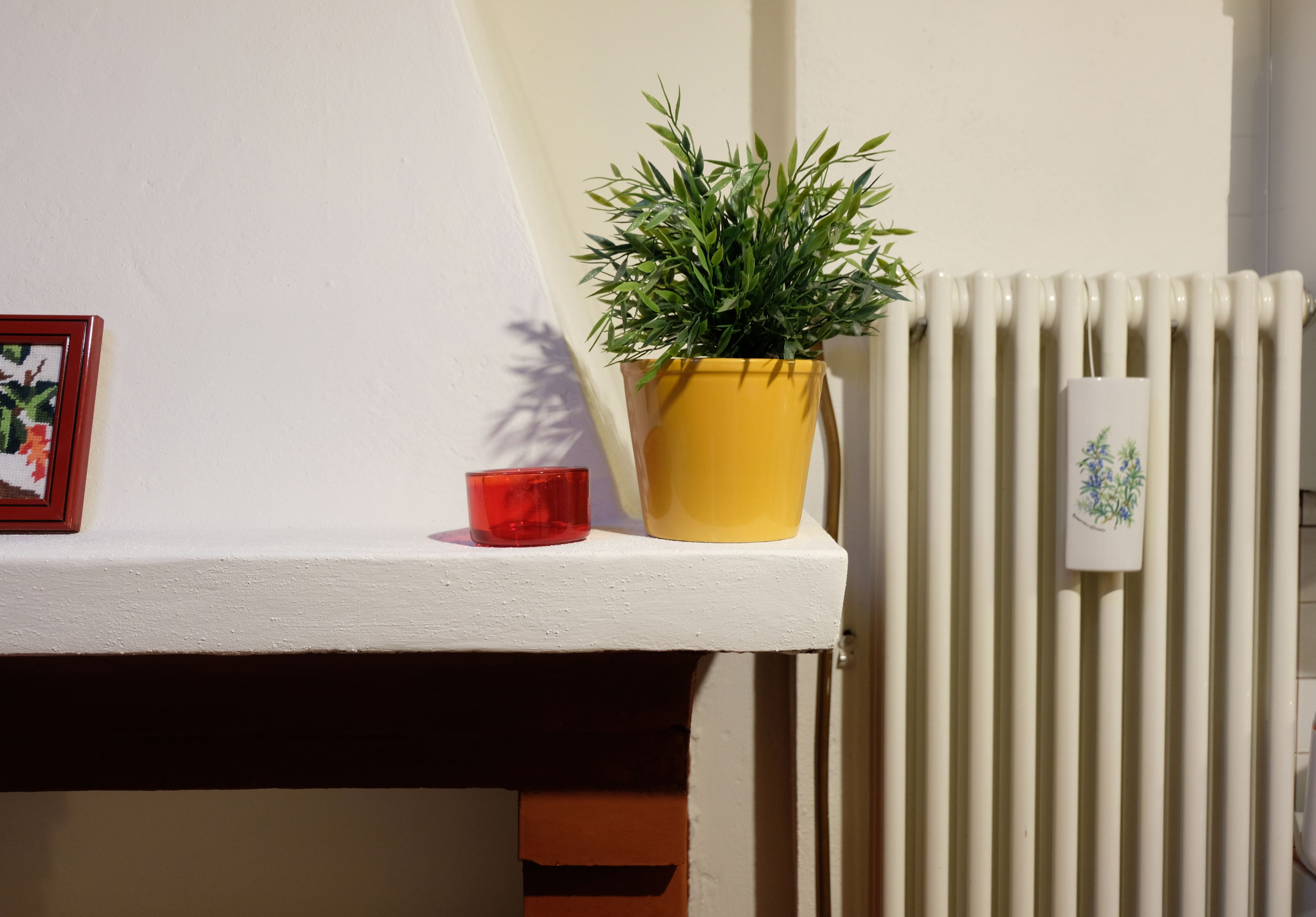 A white shelf with a yellow pot of green plant, a red candle, and a framed picture.