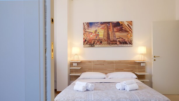 A hotel room with a bed, two bedside lamps, a painting of a cityscape, and neatly folded towels on the bed. - Marconi Airport Cozy Dream Flat (Bologna)