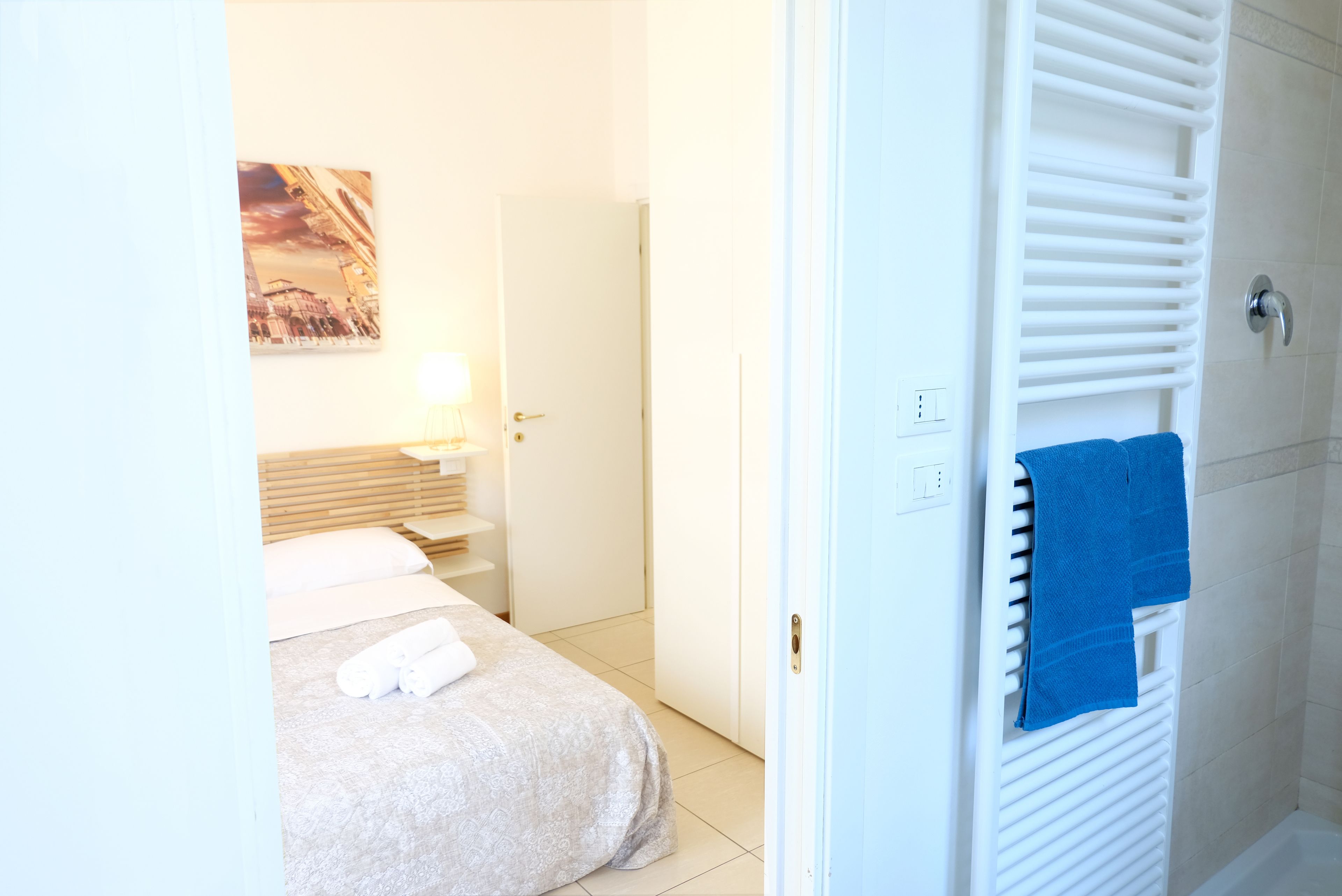 Hotellihuoneessa on sänky, yöpöytä, lamppu ja sininen pyyhe patterin päällä.