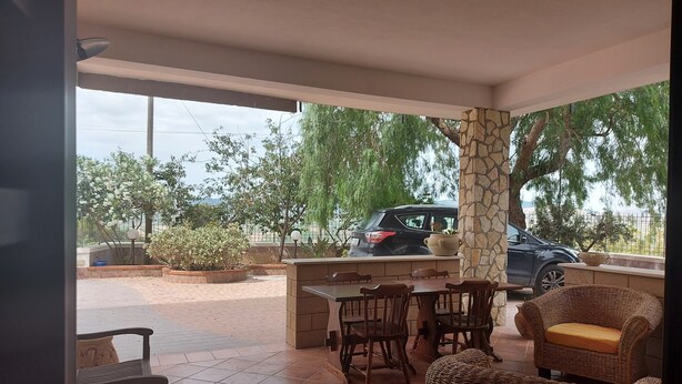 A covered outdoor seating area with a table and chairs, a stone pillar, and a view of parked cars and greenery. - Costapicca B&B (Alcamo)