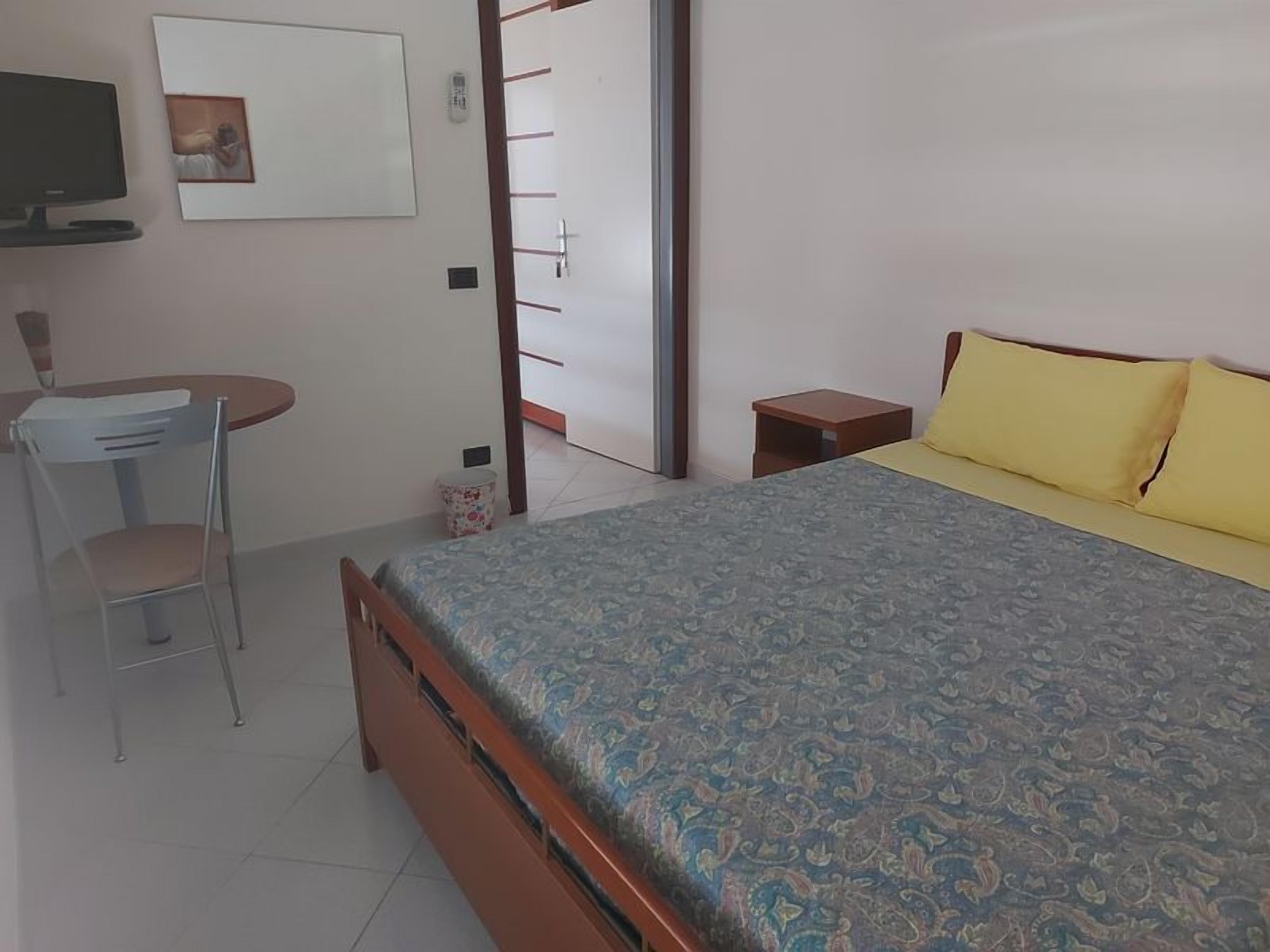 Un dormitorio con cama, una silla, una mesita, televisor y una puerta.