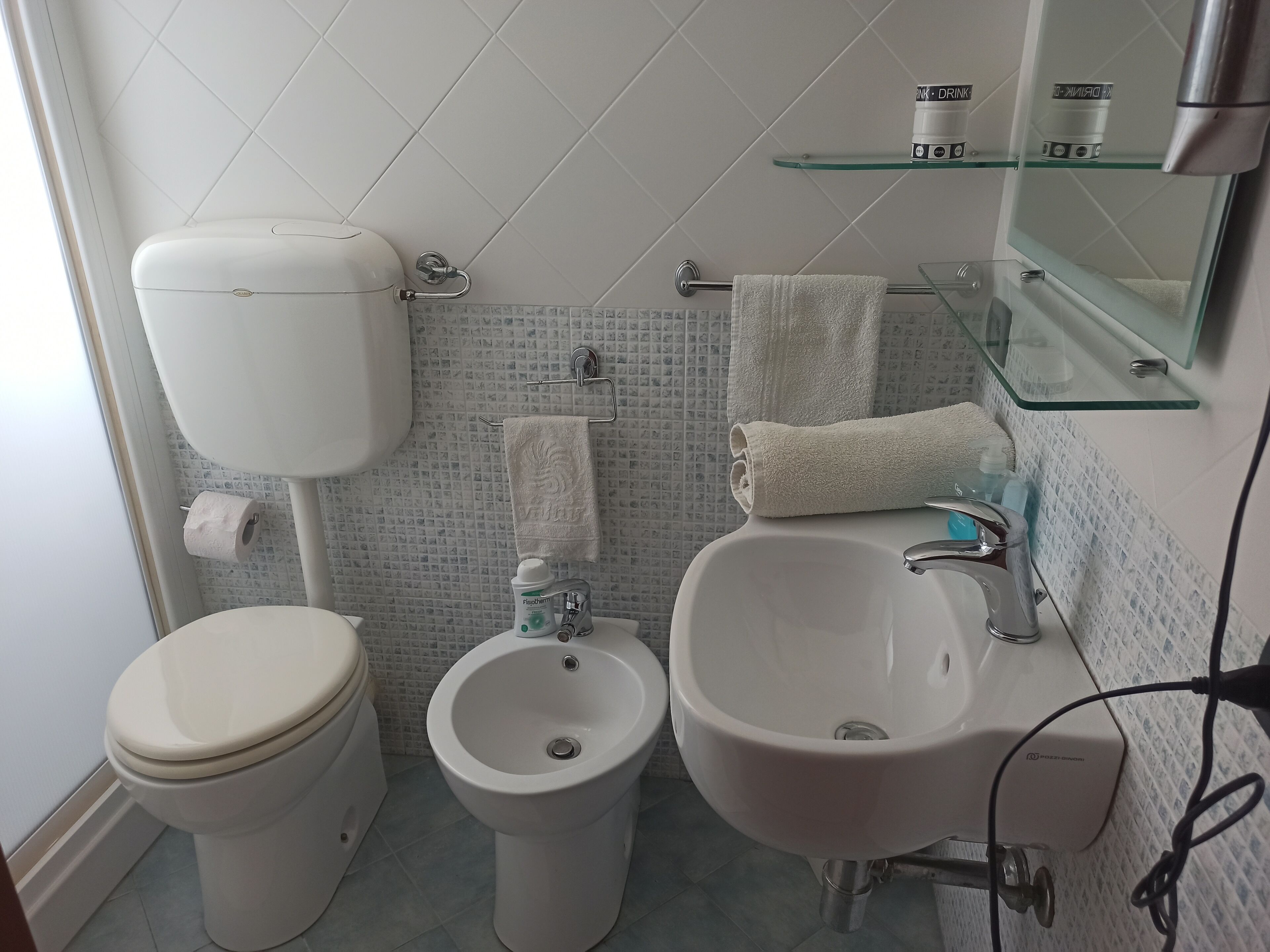 Un baño con inodoro, bidé y lavamanos, con un estante de vidrio sobre el lavamanos y un espejo en la pared.
