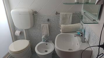 Un baño con inodoro, bidé y lavamanos, con un estante de vidrio sobre el lavamanos y un espejo en la pared.