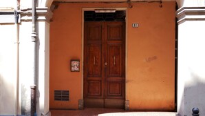 Une porte en bois à deux battants, surmontée d’un portail en métal, est intégrée à un bâtiment dont la façade est peinte en orange et au-dessus de laquelle on peut voir une petite enseigne.