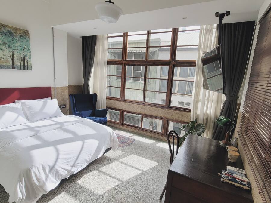 Hotellihuone, jossa on sänky, työpöytä lampun kanssa, tuoli, ikkuna verhoilla ja taulu seinällä.