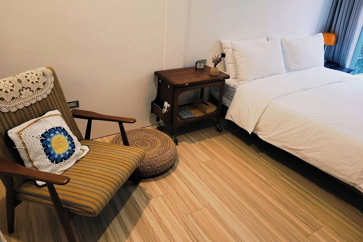 Habitación de hotel con una silla de madera, una mesita de noche pequeña de madera, una cama con ropa blanca y una ventana con cortinas.