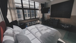 Ein Hotelzimmer mit Bett, Schreibtisch, Stuhl, Fernseher und Fenster mit Blick auf Gebäude.